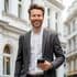 Immobilienprofi in einem grauen Anzug und weißem Hemd lächelt selbstbewusst, während er ein Smartphone hält und vor klassischen weißen Gebäuden mit Bogenfenstern und eleganten architektonischen Details steht.