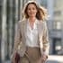 Professionelle Frau in einem beigefarbenen Business-Anzug und einer weißen Bluse, die selbstbewusst in einer städtischen Umgebung geht. Ihr Haar wird vom Wind verweht und sie trägt ein Ledermäppchen, während der Hintergrund eine verschwommene Stadtansicht zeigt.