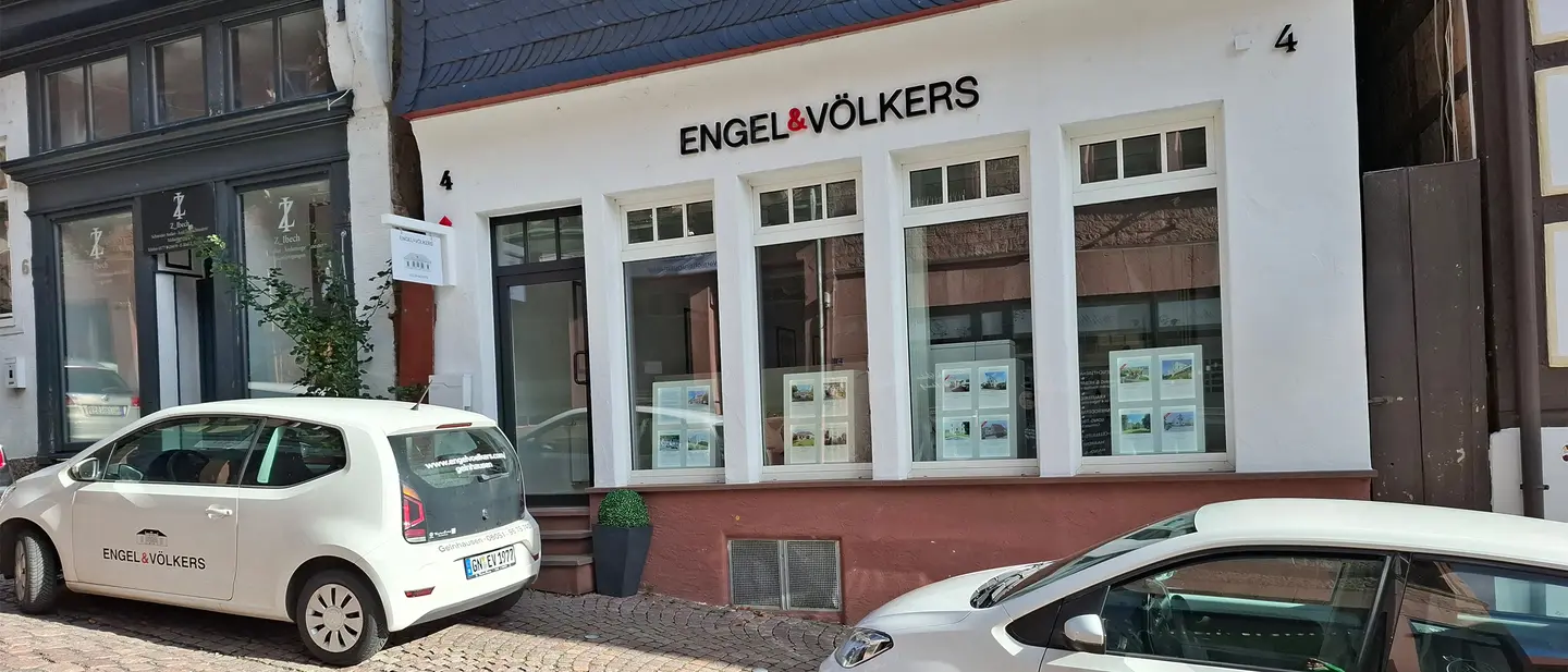 Engel & Völkers real estate office with white cars parked in front. The building is white with large windows displaying property listings.