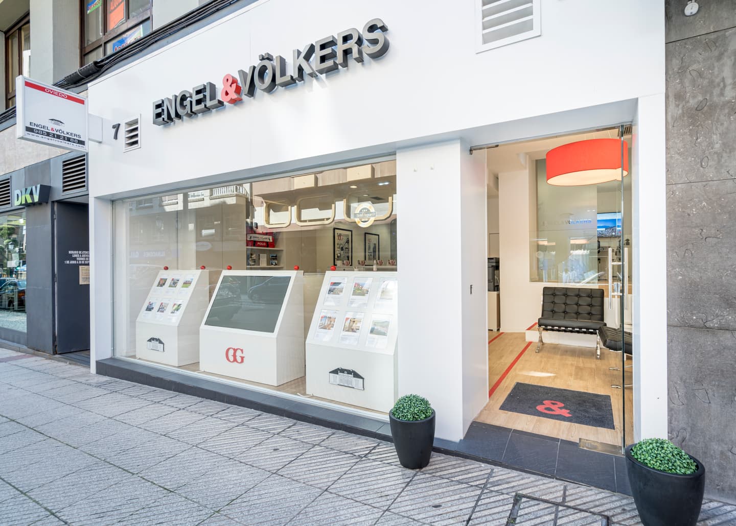 Eine Engel & Völkers Filiale mit einer Glasfassade. Weiße Displays präsentieren Immobilienangebote. Eine rote Lampe hängt im Inneren in der Nähe eines schwarzen Ledersessels.
