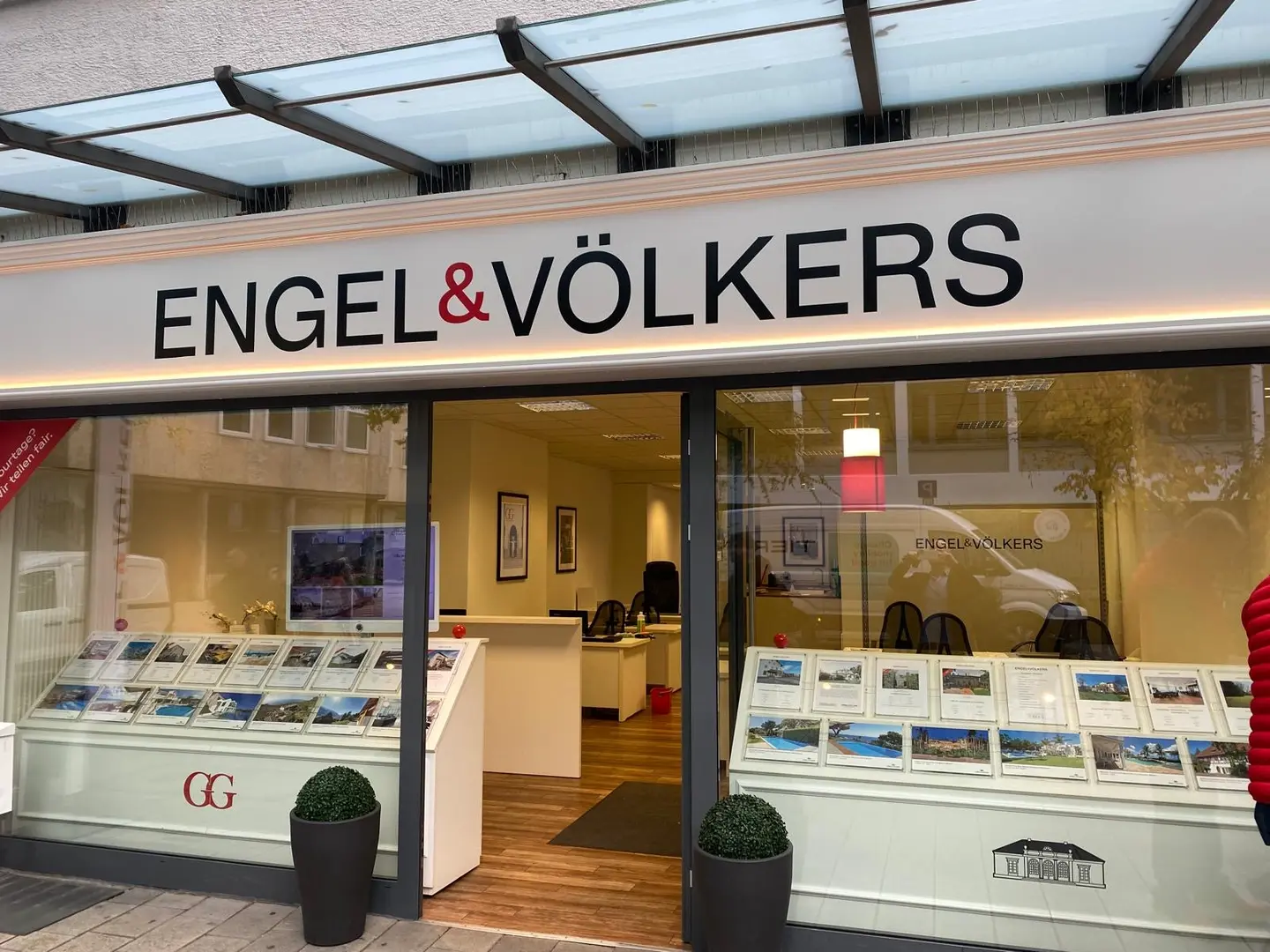 Engel & Völkers real estate office storefront with property listings displayed in the window. The logo is above the entrance.