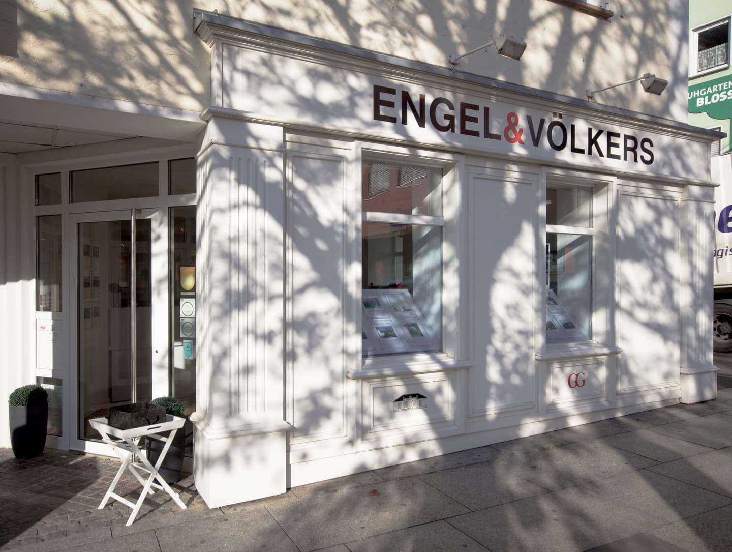 Exterior of Engel & Völkers real estate office with white trim, large windows, and tree shadows. A white folding table sits outside the entrance.