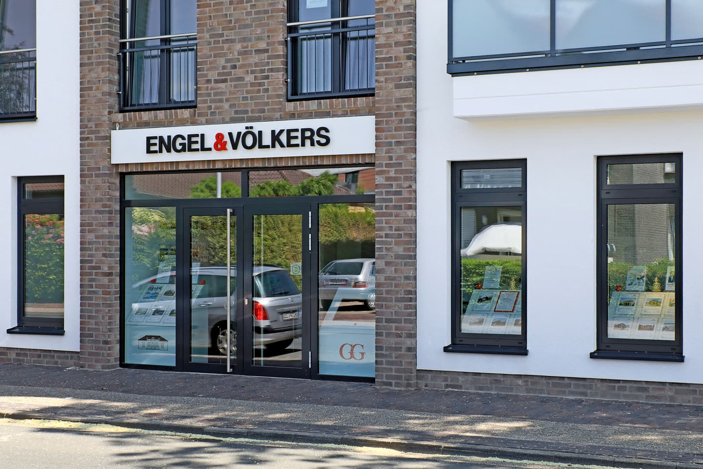 Engel & Völkers real estate office exterior. Brick and white building with black framed windows and glass doors. Cars are reflected in the glass.