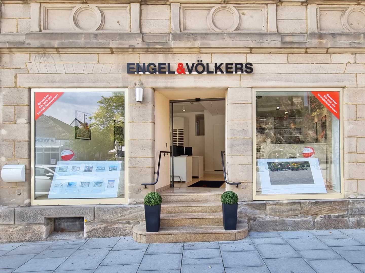 Engel & Völkers Immobilienbüro, Außenansicht. Steingebäude mit großen Fenstern, Schild über der Tür und Topfpflanzen auf den Stufen.
