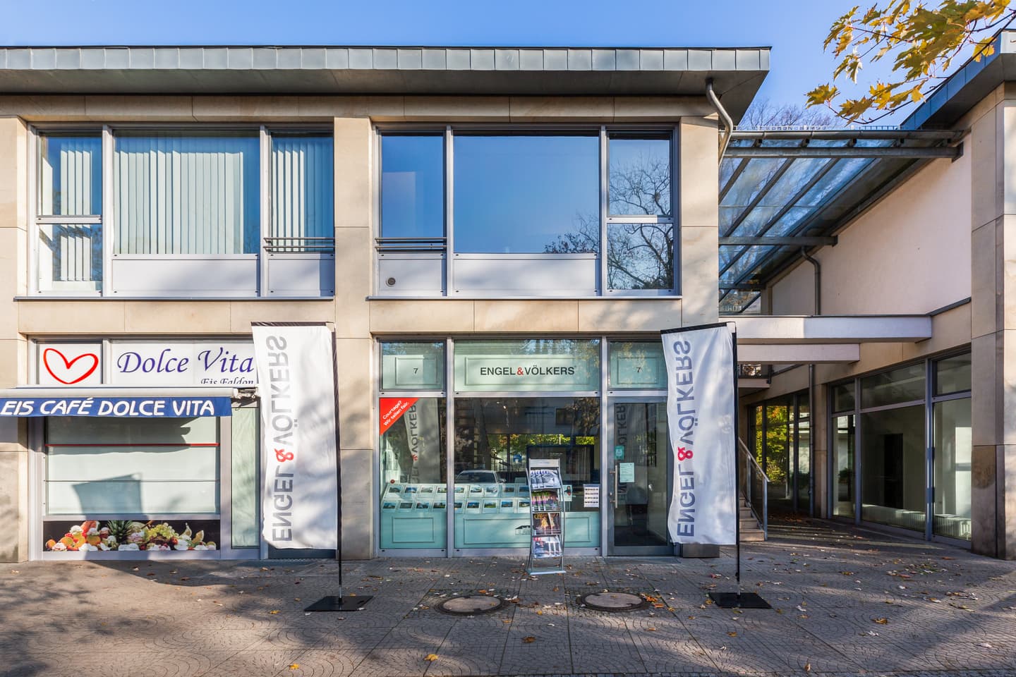 Engel & Völkers office storefront with large windows and flags on a sunny day. Next door is a cafe called Dolce Vita.
