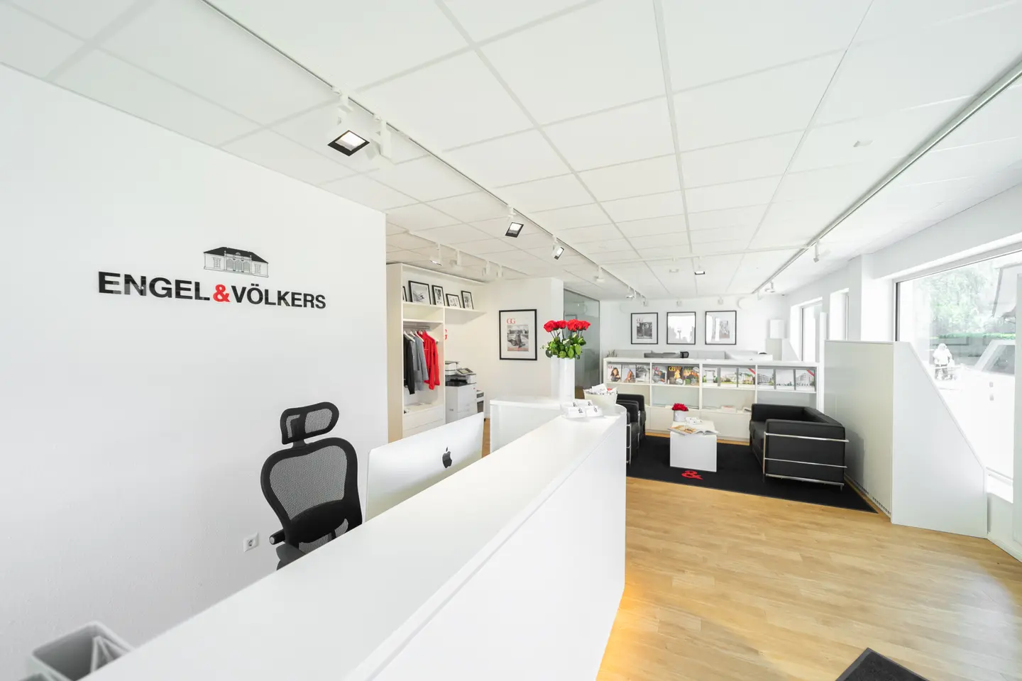 Engel & Völkers office interior. White walls, light wood floors, black sofa, and a white reception desk with the company logo.