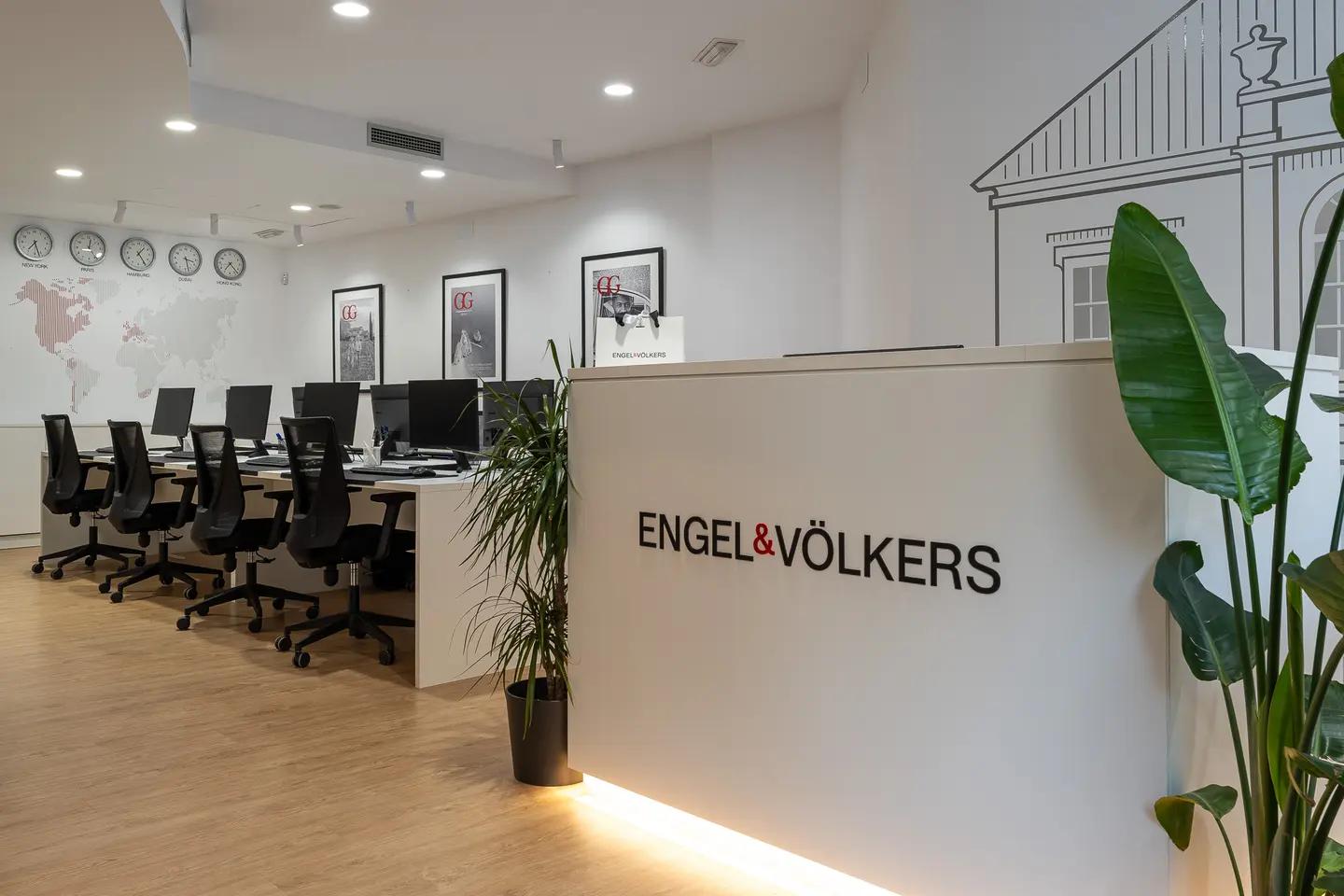 Engel & Völkers office interior. White reception desk with logo, world map, clocks, desks with computers, and potted plants.