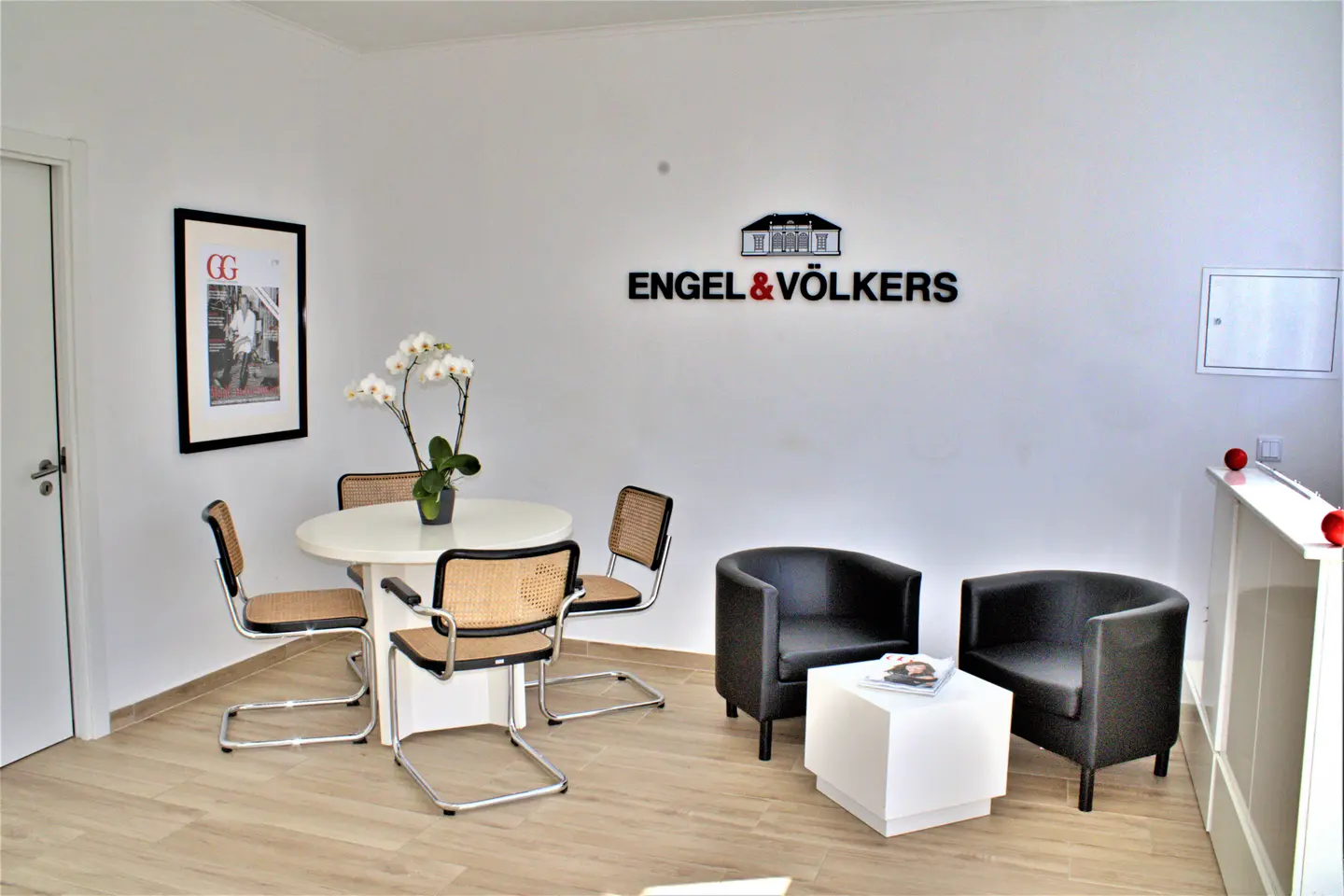 Engel & Völkers office interior with a white table, four chairs, two black armchairs, and the company logo on the wall.