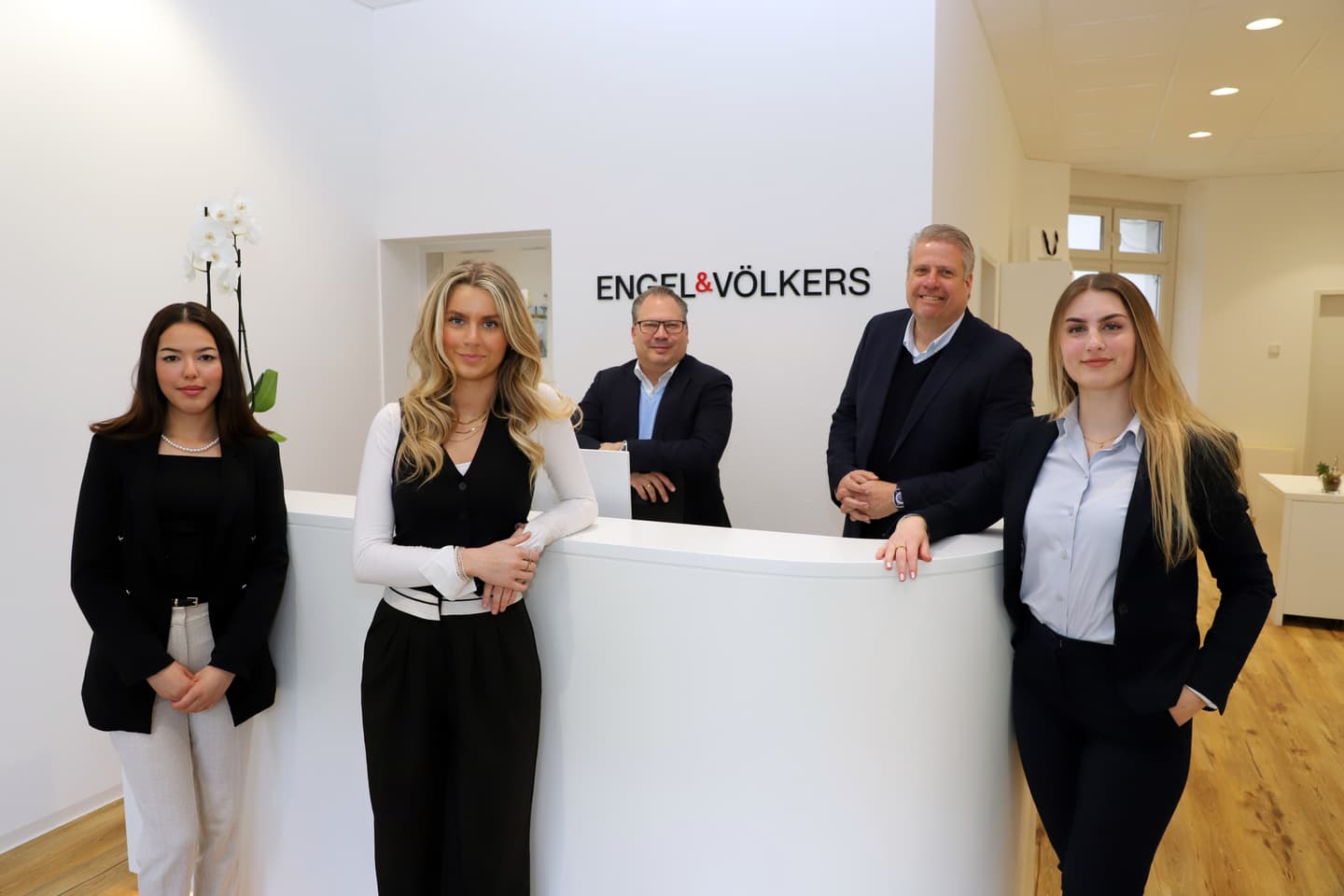 Engel & Völkers real estate team of four poses behind a white reception desk in a modern office with the company logo on the wall.