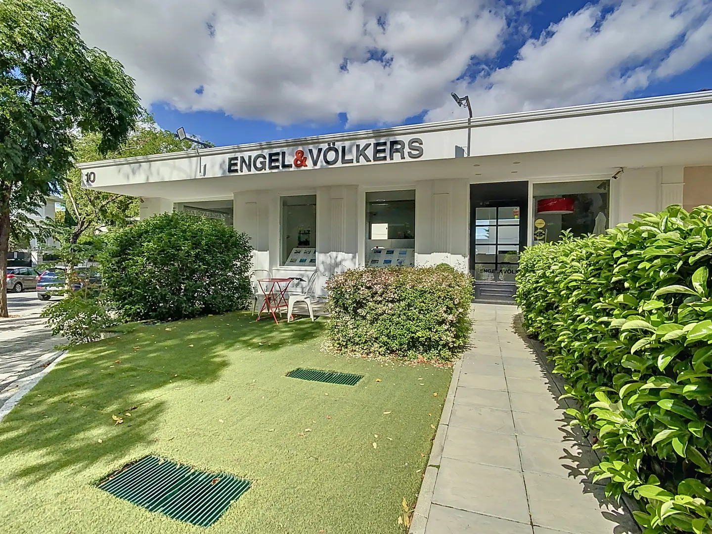 Engel & Völkers Immobilienbüro mit einer grünen Rasenfläche, Büschen und einem roten Tisch mit Stühlen.