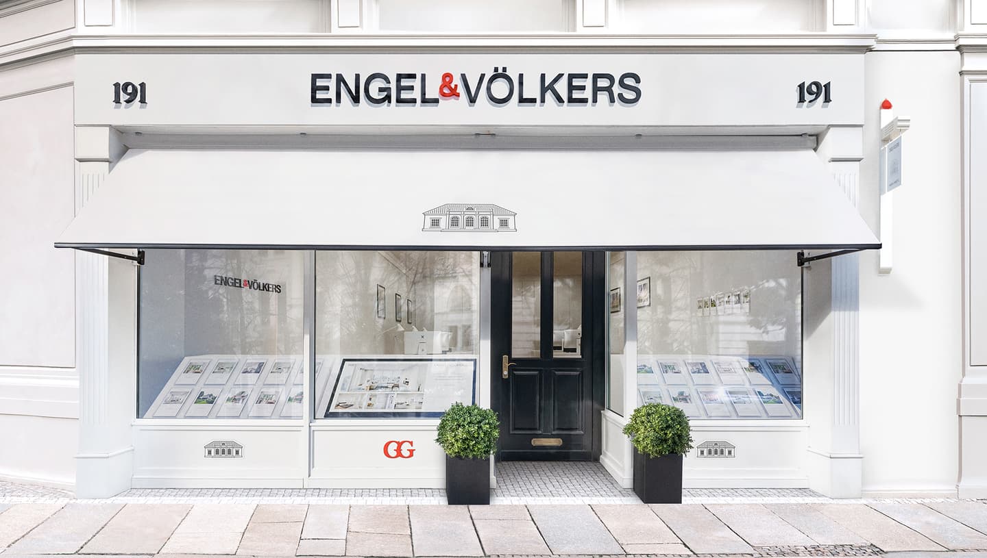 Engel & Völkers real estate storefront with white awning, black door, and property listings displayed in the window.