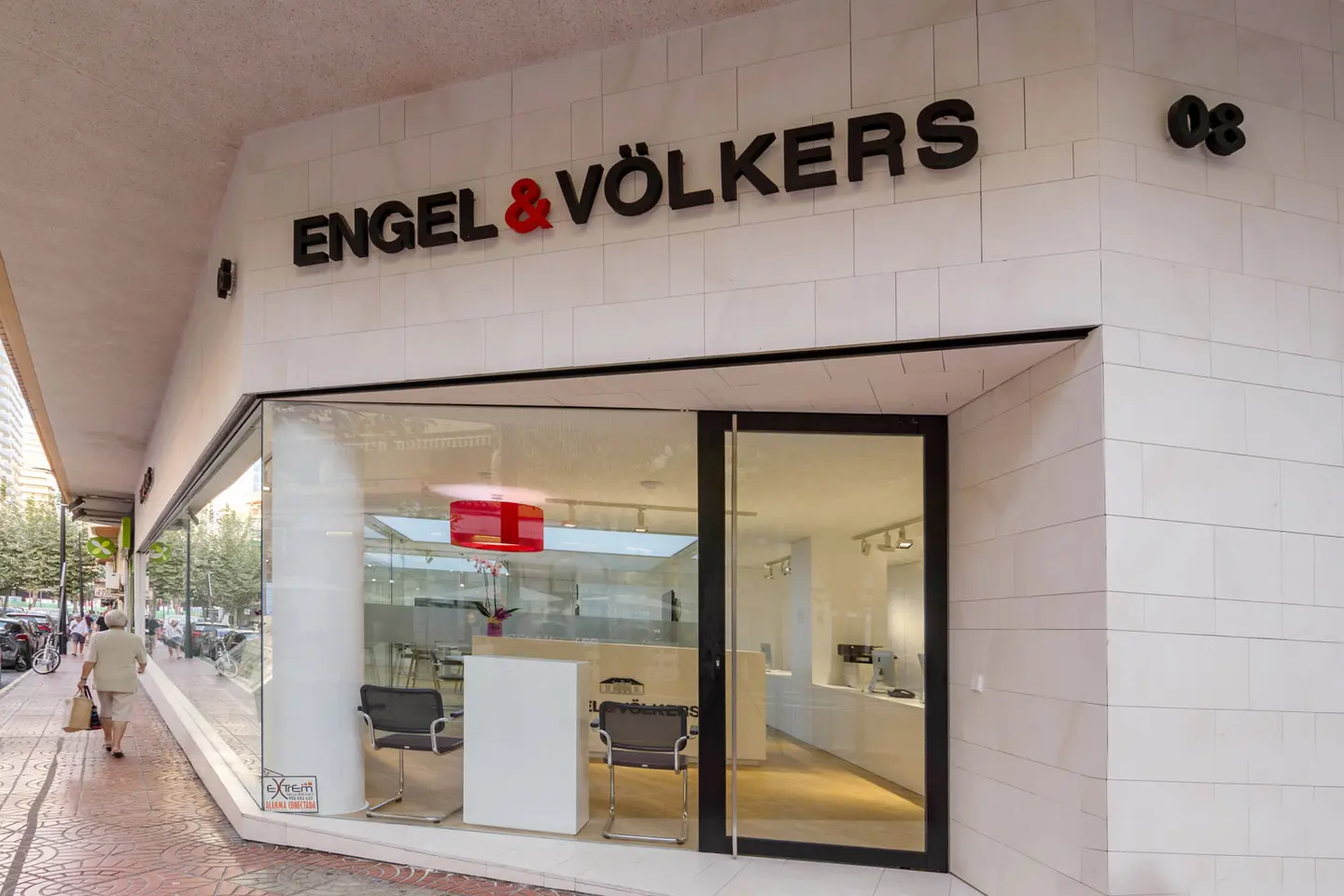 Engel & Völkers Immobilienbüro, Außenansicht. Schwarzes Logo auf weißen Ziegeln. Glasfassade mit Stühlen und einer roten Lampe im Inneren.