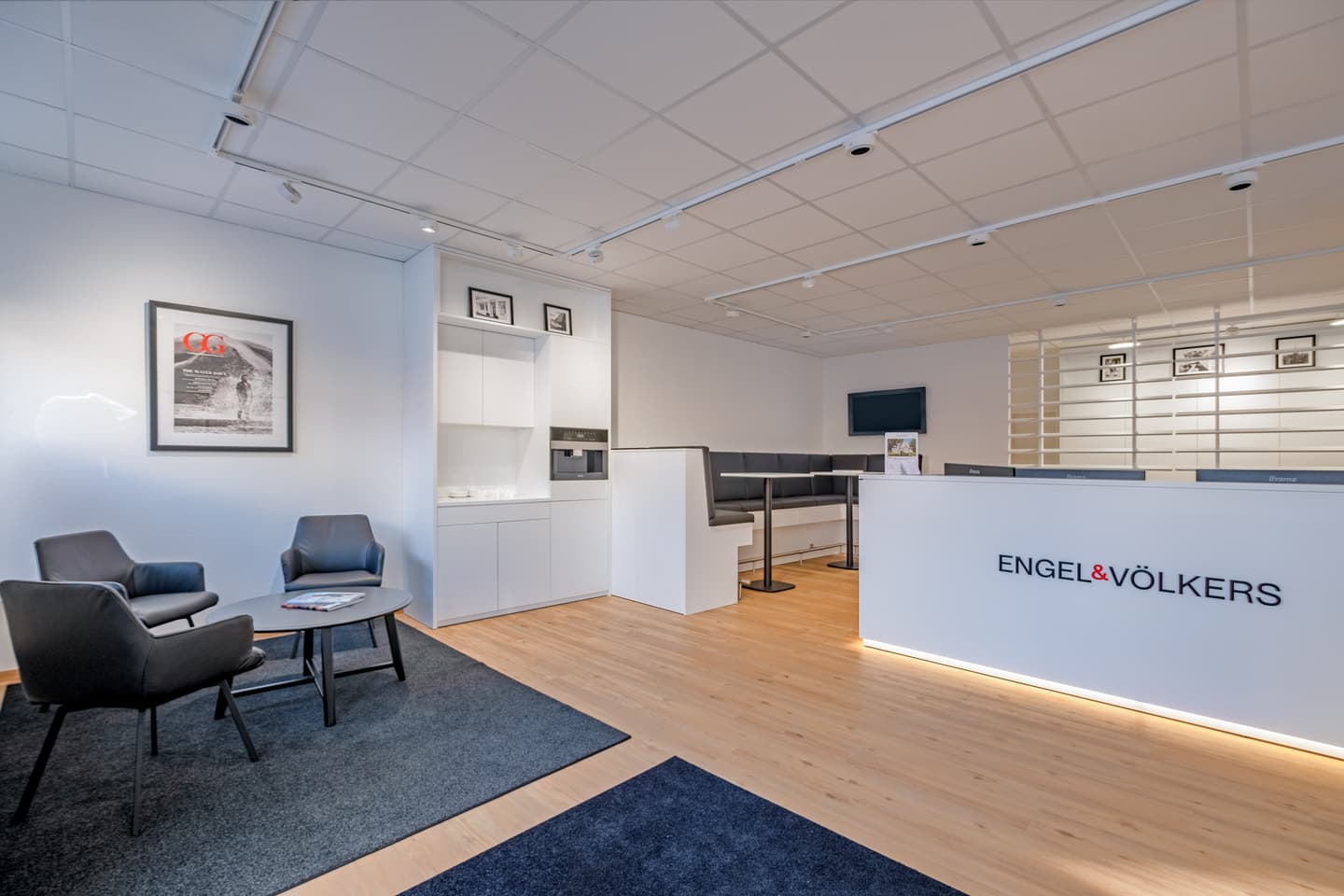 Engel & Völkers office interior with gray chairs, a coffee table, and a white reception desk with the company logo.