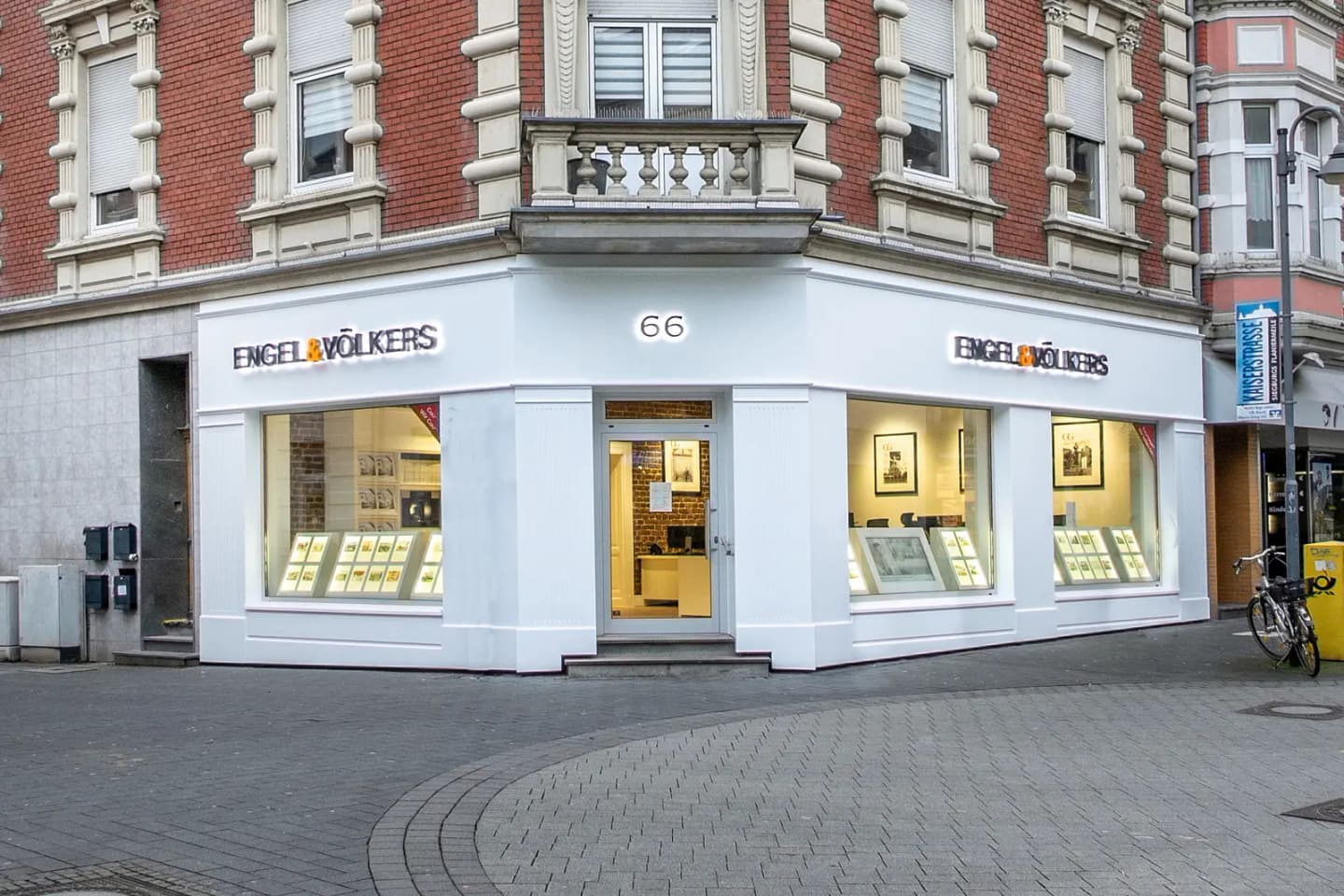 Engel & Völkers real estate office on a corner. The building is brick with white trim and large windows displaying framed photos. A bicycle is parked nearby.
