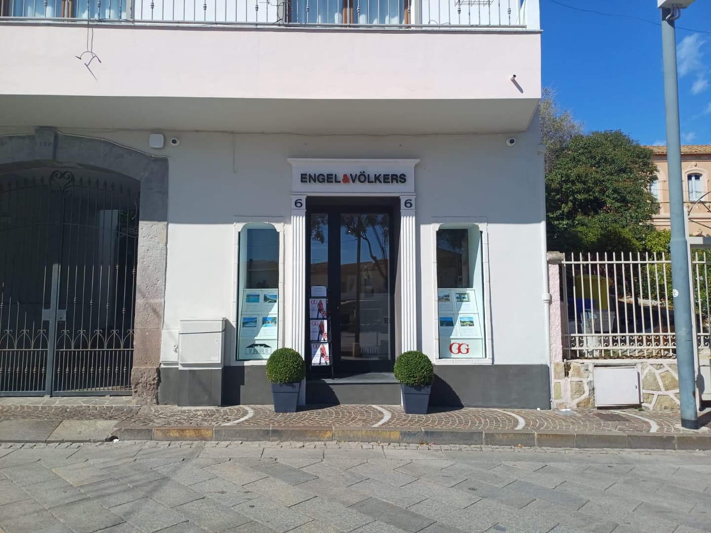 Engel & Völkers real estate office with a white facade, black door, and window displays. Two potted plants flank the entrance.