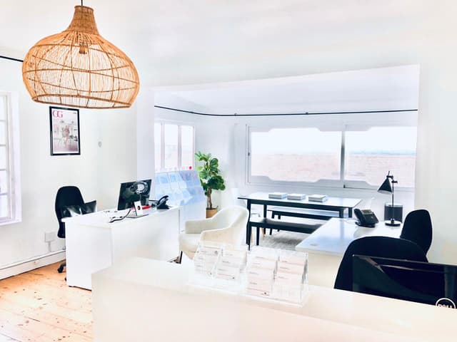 Bright real estate office with white walls, light wood floors, and a large window with an ocean view. A woven pendant light hangs above the reception desk.