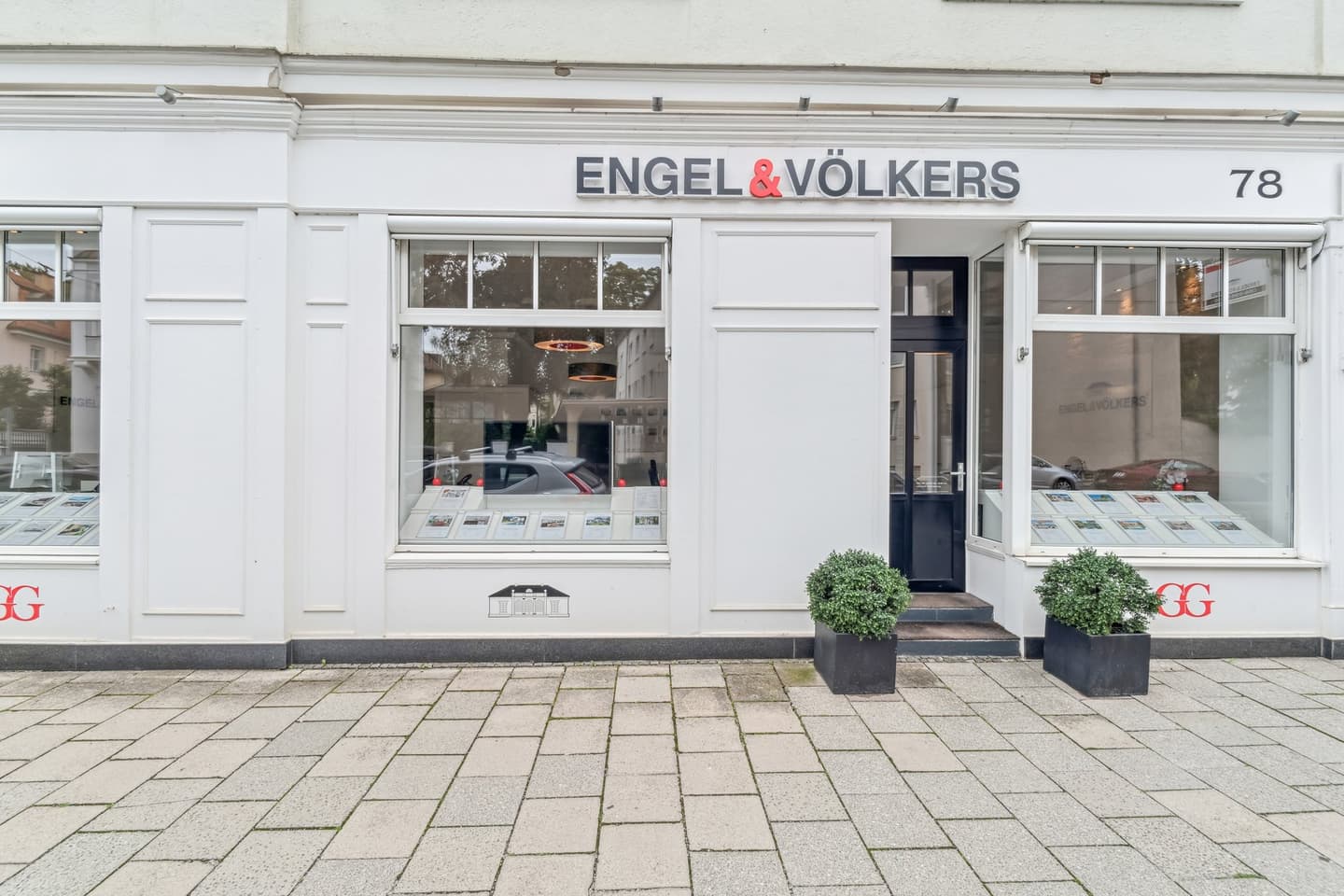 Engel & Völkers real estate office with white facade, large windows displaying property listings, and two potted plants flanking the entrance.