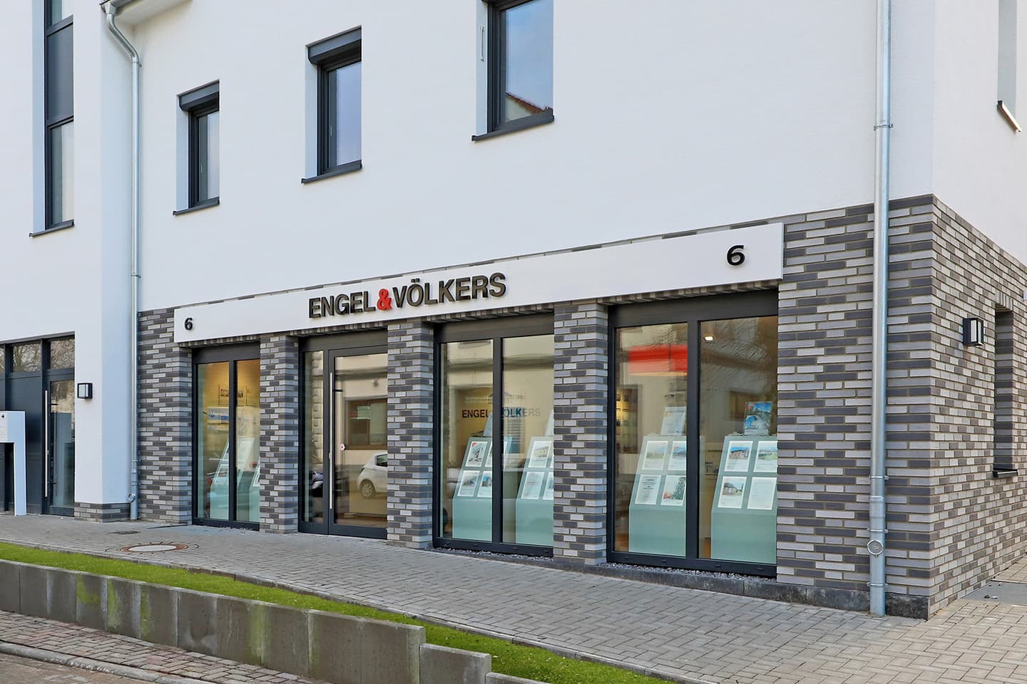 Engel & Völkers Immobilienbüro mit grauen Ziegeln und großen Fenstern, in denen Immobilienangebote ausgestellt sind.
