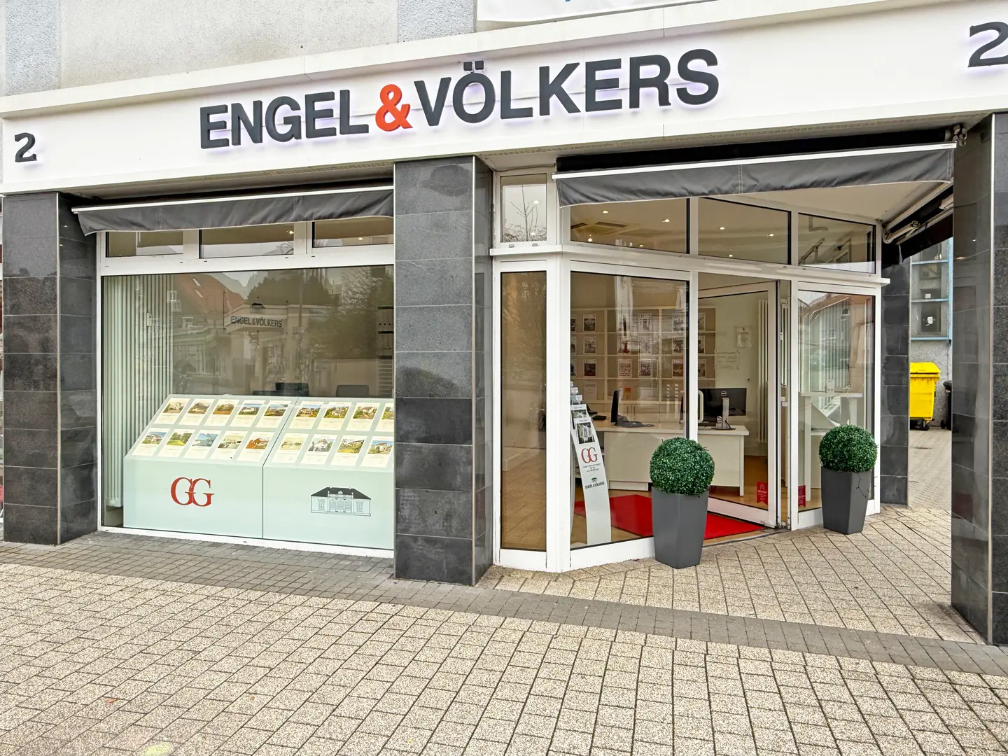 Exterior of Engel & Völkers real estate office with a display of property listings in the window. The entrance is open, flanked by potted plants.