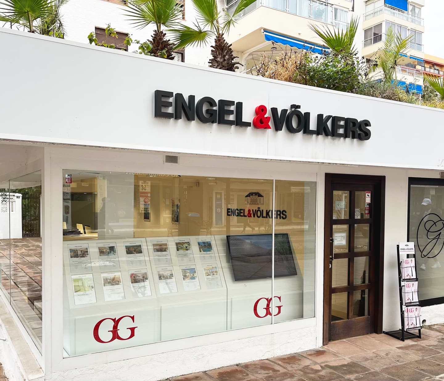 Engel & Völkers real estate office with a white facade, black lettering, and a red ampersand. Palm trees are visible above the building.