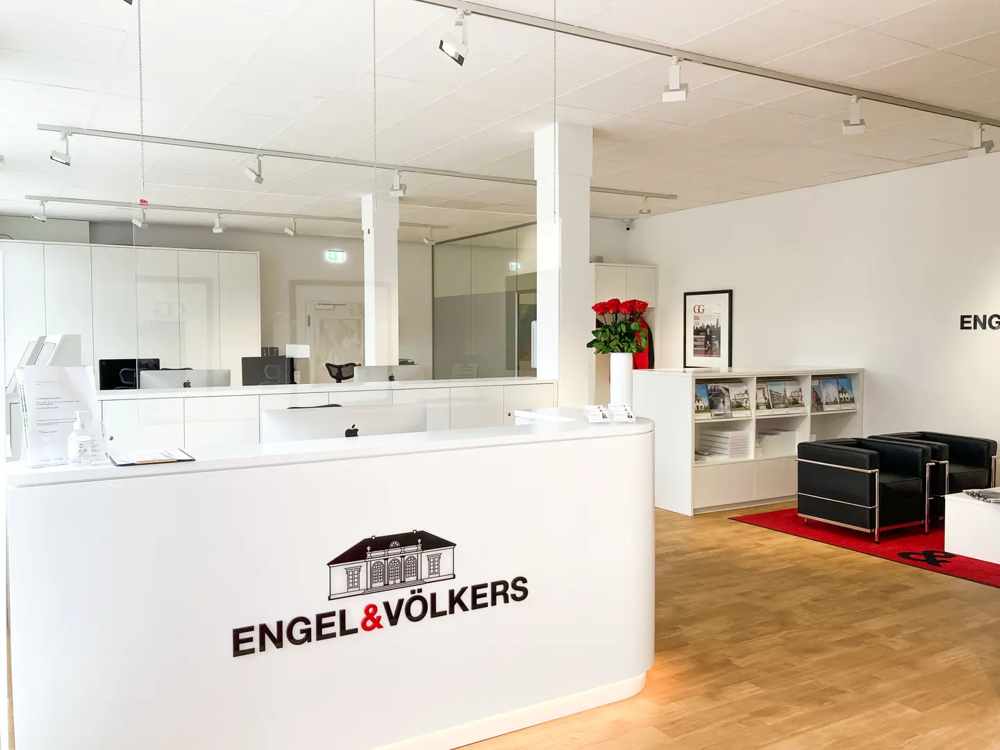 Helles Interieur eines Immobilienbüros mit weißem Empfangstresen, Engel & Völkers Logo und Sitzecke mit schwarzen Stühlen auf einem roten Teppich.
