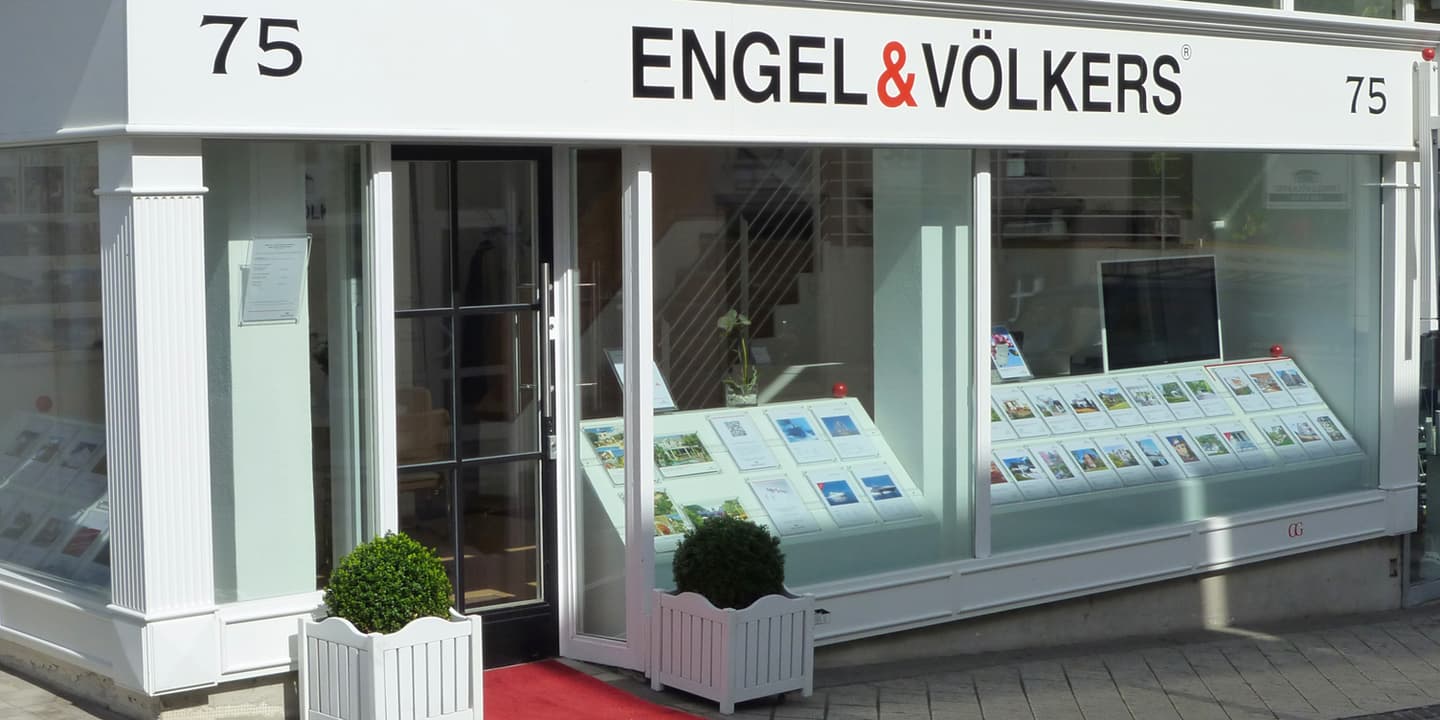 Engel & Völkers real estate office exterior. White building with glass windows displaying property listings. Red carpet leads to the door.