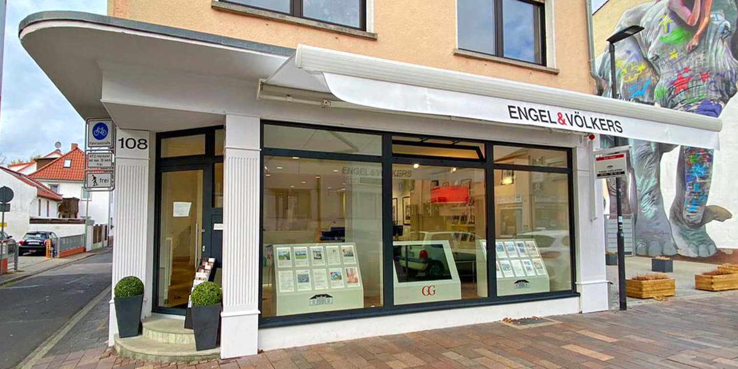 Engel & Völkers Immobilienbüro mit großen Fenstern, in denen Immobilienangebote ausgestellt sind. An der Seite des Gebäudes befindet sich ein Elefanten-Wandbild.