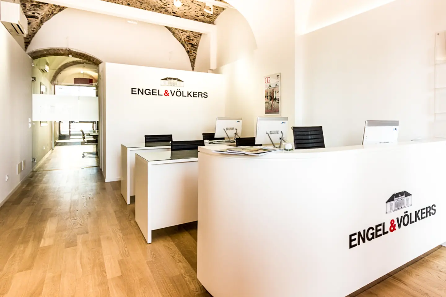 Engel & Völkers Immobilienbüro-Interieur. Weiße Wände, Holzböden, Empfangstresen mit Logo, Computer und schwarze Stühle.
