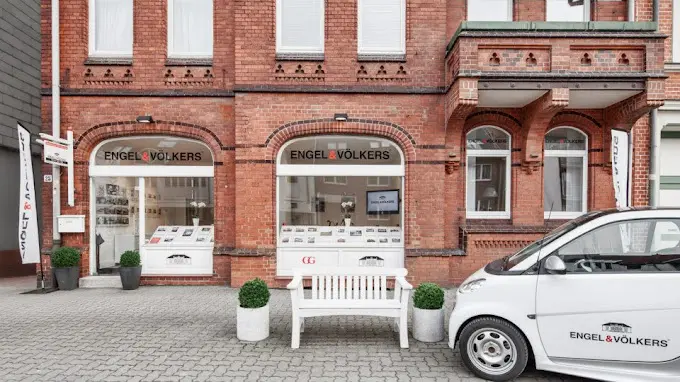 Engel & Völkers Immobilienbüro in einem roten Backsteingebäude mit einer weißen Bank und einem gebrandeten Smart, der draußen geparkt ist.