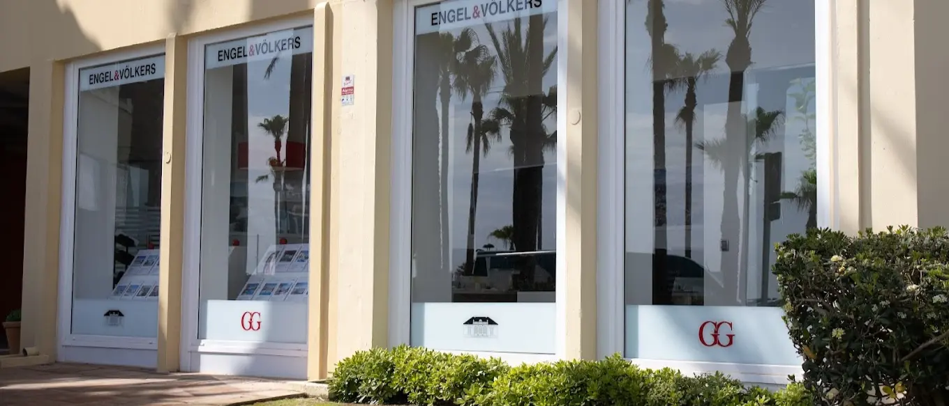 Oficina immobiliària d'Engel & Völkers amb grans finestrals que reflecteixen palmeres i mostren llistats de propietats.