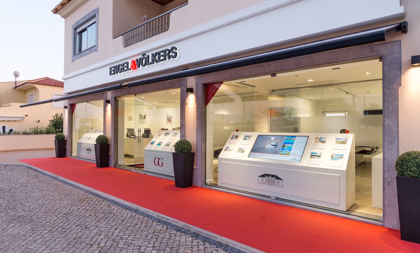 Engel & Völkers real estate office with a red carpet entrance. Displays of properties are visible through the glass windows.