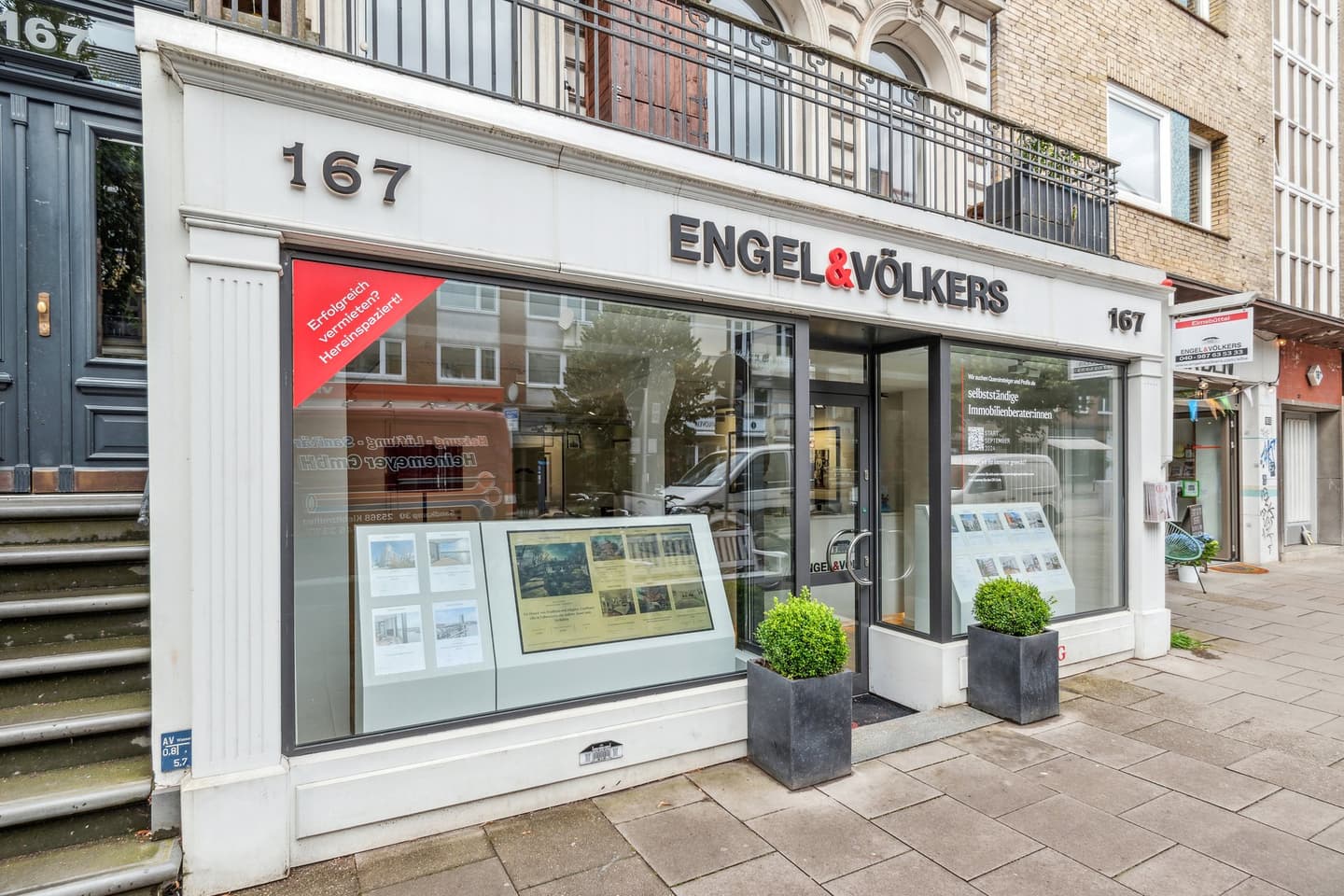 Engel & Völkers real estate office storefront with large windows, displays, and potted plants. The building is brick and white.