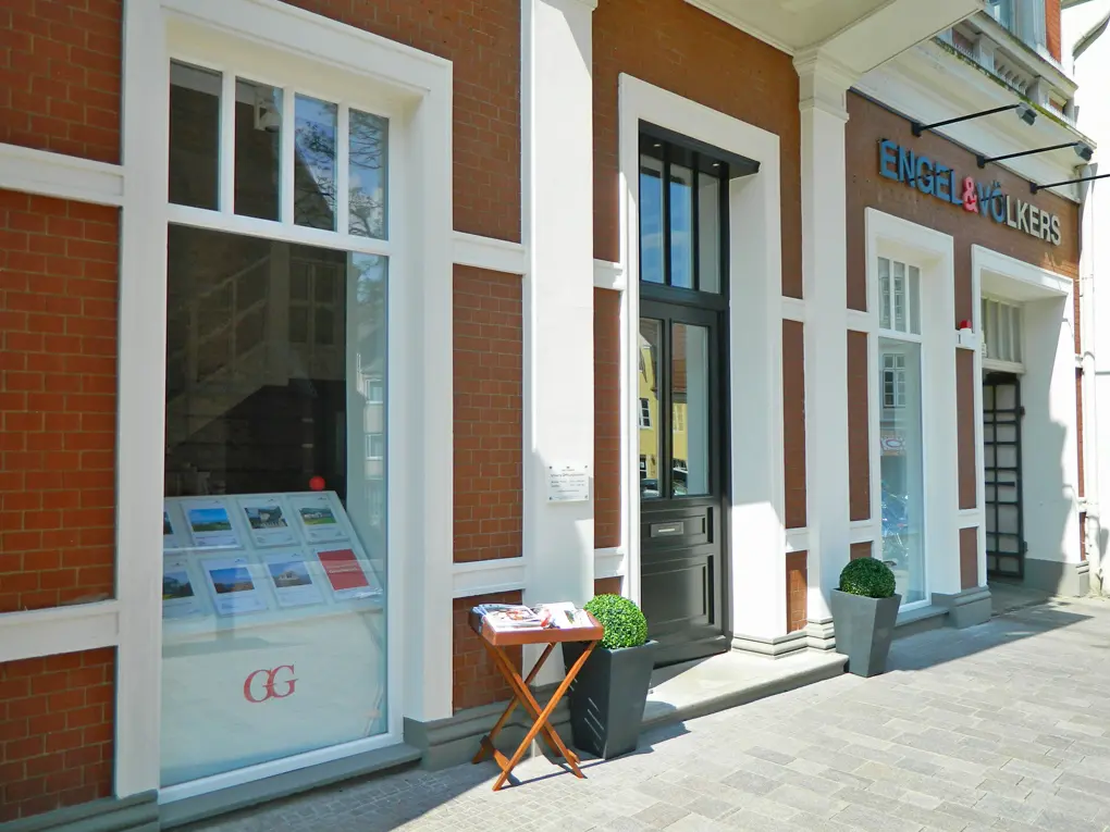Außenansicht des Engel & Völkers Immobilienbüros mit Backsteinfassade, weißen Zierelementen, schwarzer Tür und Fenster mit Immobiliendisplays.