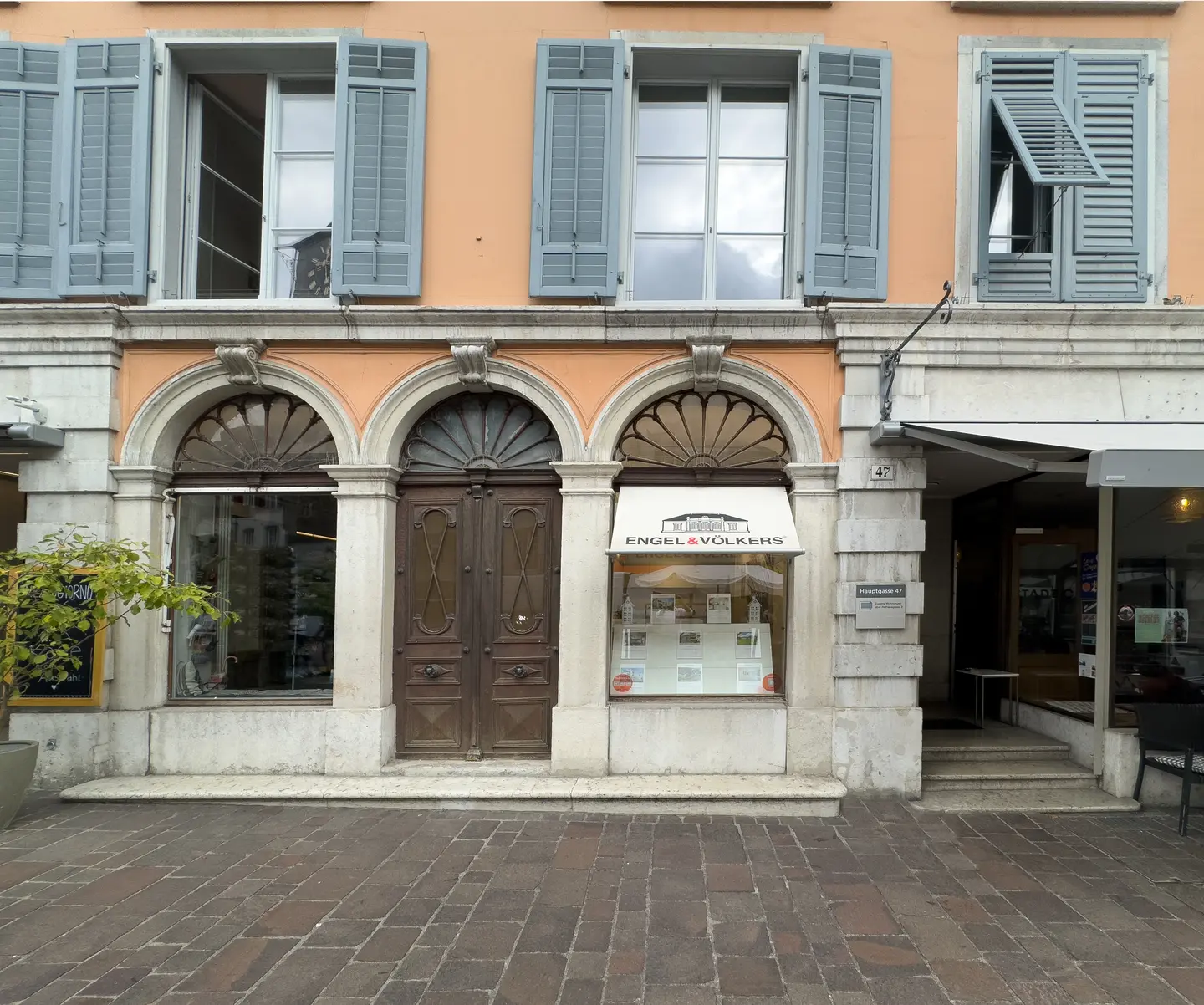 Esterno di un ufficio immobiliare Engel & Völkers con finestre ad arco, porte marroni e persiane blu su un edificio arancione.