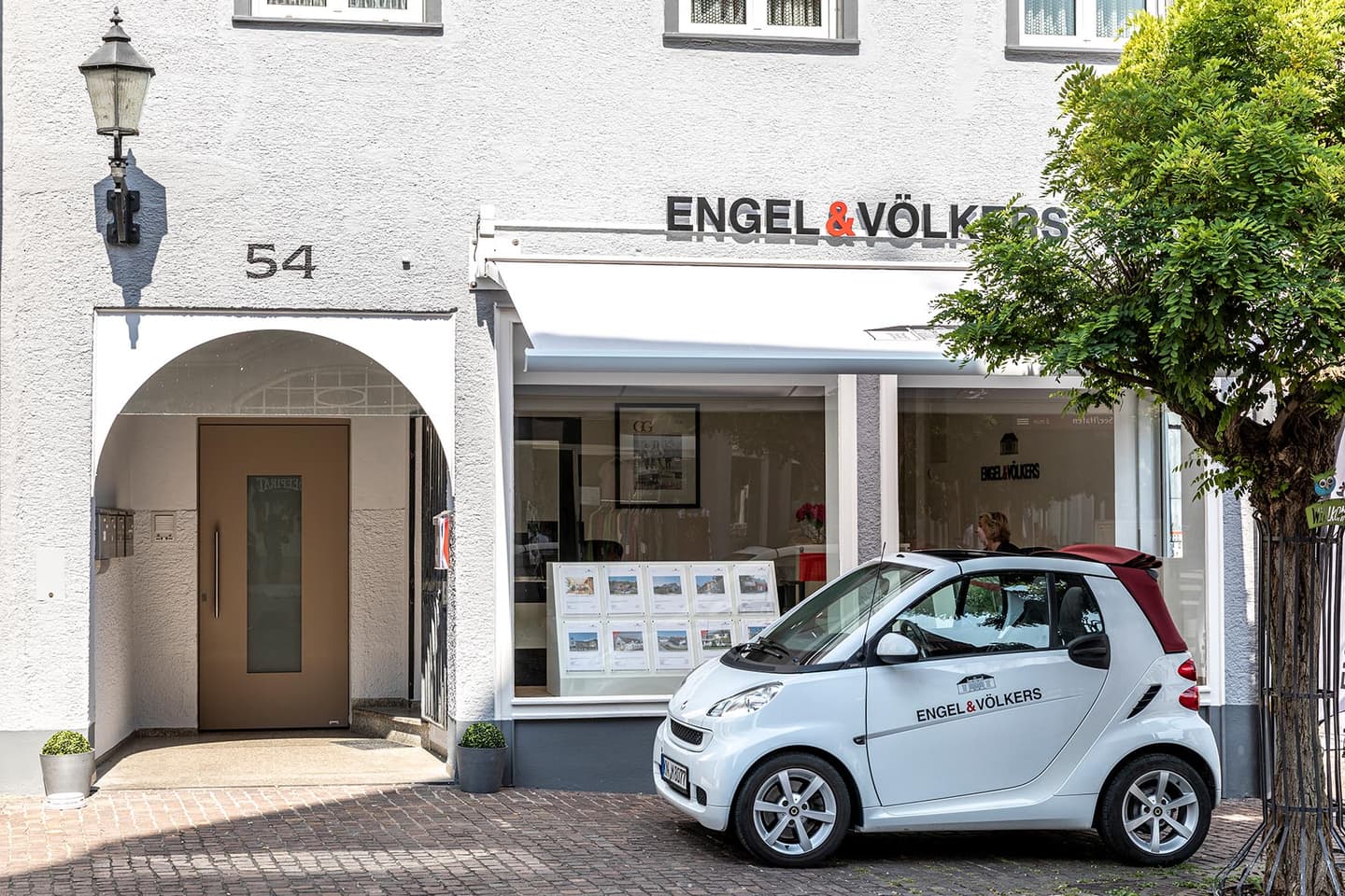 Engel & Völkers office exterior. White building with "54" above arched doorway. White car with logo parked outside.