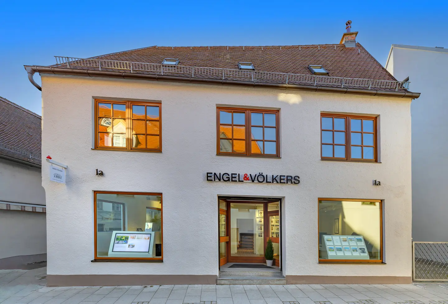 Engel & Völkers real estate office. A white building with brown framed windows and a brown tiled roof. The company name is above the open front door.