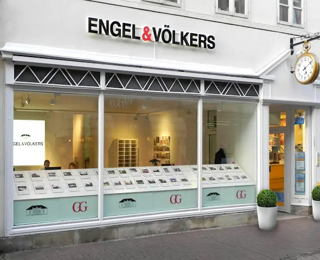 Engel & Völkers real estate office storefront with property listings in the window. A clock hangs outside the entrance.