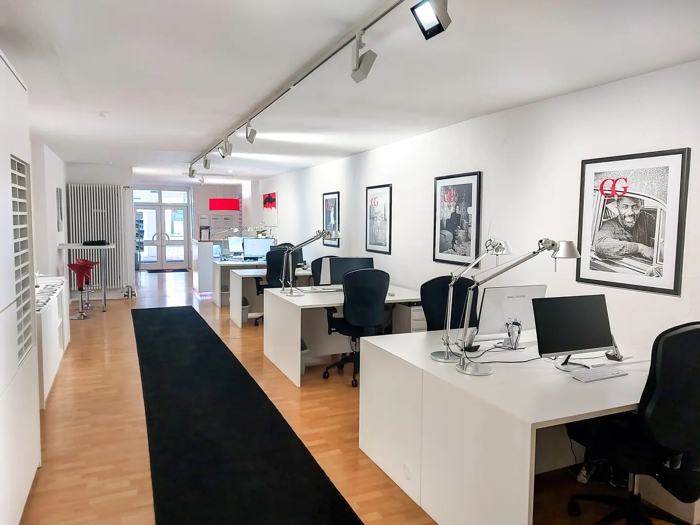 Bright office space with white walls, wood floors, and black accents. Desks with computers line the room, illuminated by track lighting. Framed art adorns the walls.