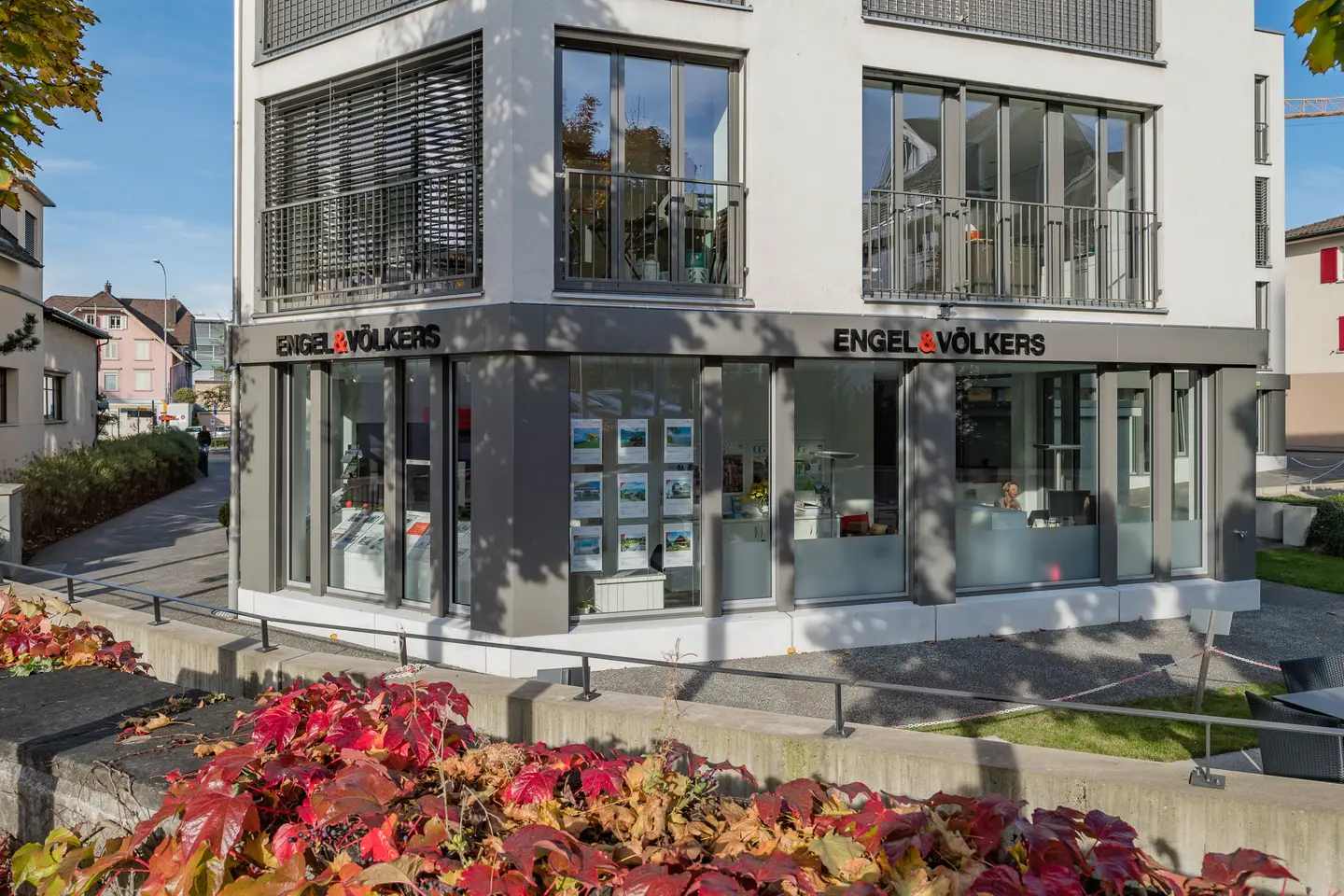 Agence immobilière Engel & Völkers avec de grandes fenêtres présentant des annonces immobilières. Du lierre rouge grimpe sur le mur au premier plan.