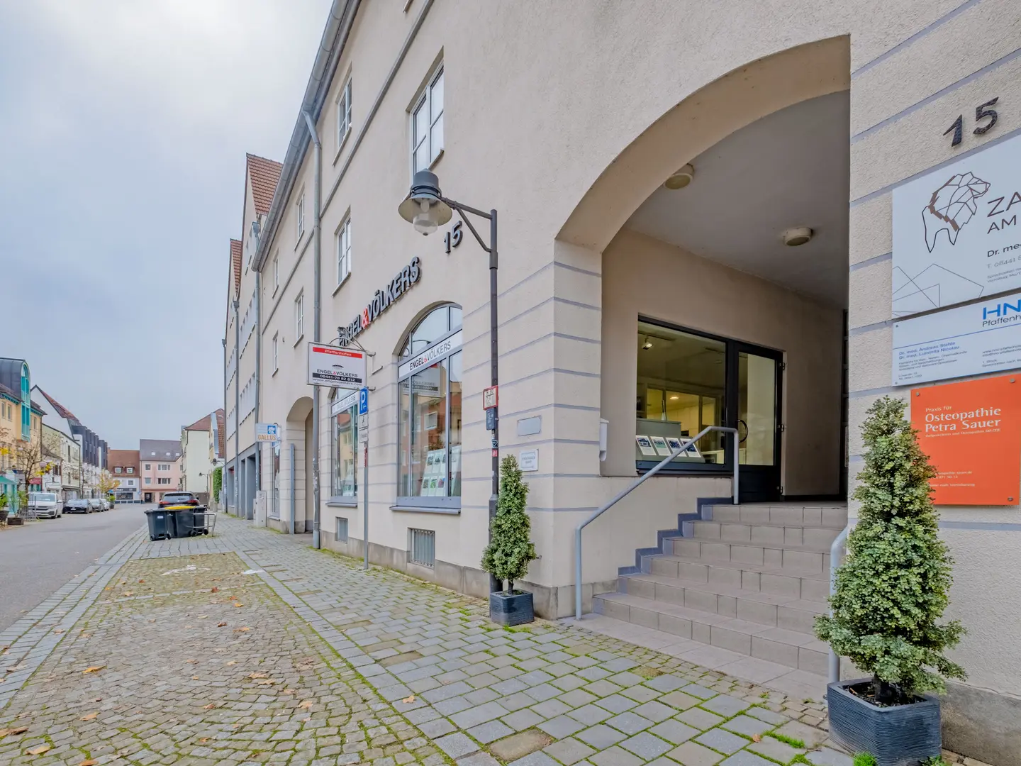 Building exterior with "Engel & Völkers" sign above a window. A stone walkway leads to an arched entrance with stairs. Signage is visible.