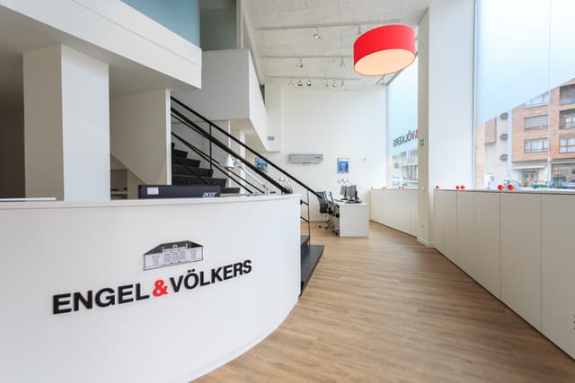 Oficina de Engel & Völkers. Mostrador de recepción blanco con logotipo, escaleras negras, piso de madera y una lámpara colgante roja.