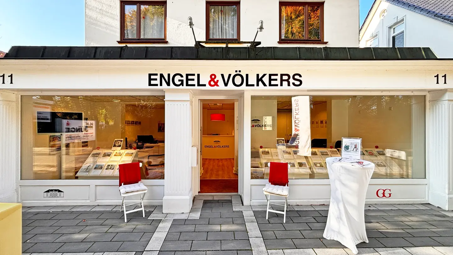 Engel & Völkers real estate office with white facade, large windows displaying property listings, and two chairs with red throws.