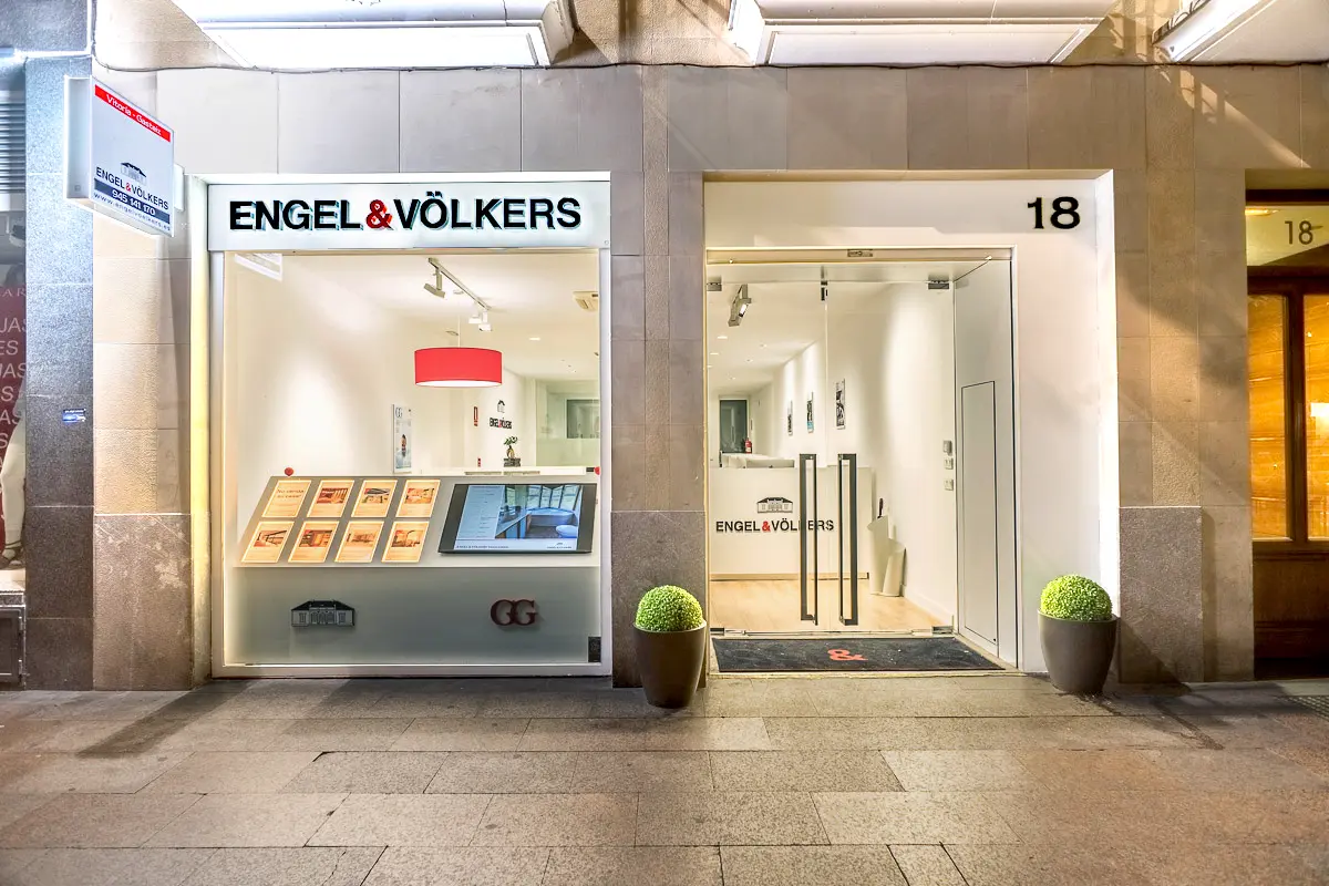 Engel & Völkers real estate storefront with a glass door and display window. Potted plants flank the entrance. The address is marked as 18.