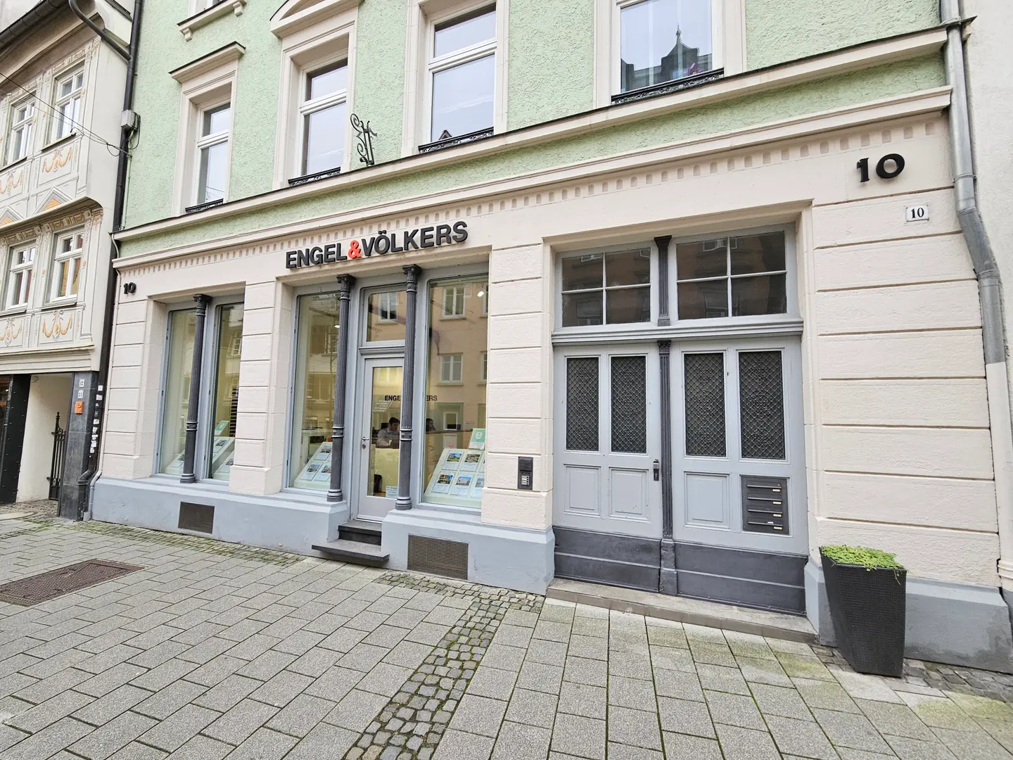 Außenansicht des Engel & Völkers Immobilienbüros mit großen Fenstern, einer grauen Tür und der Nummer 10 am Gebäude.