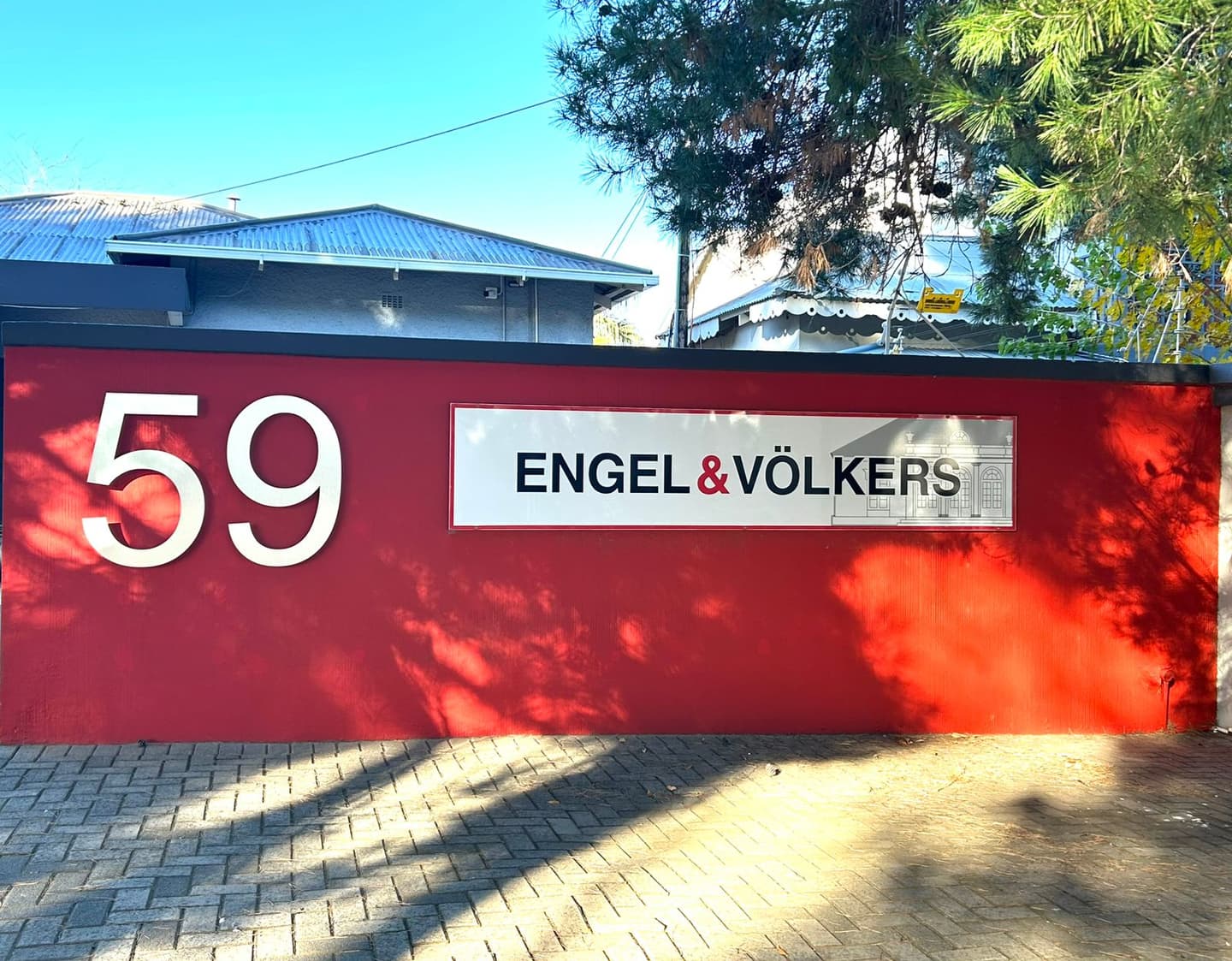 Red wall with "59" and "Engel & Völkers" sign. Shadows of trees on the wall and brick walkway.