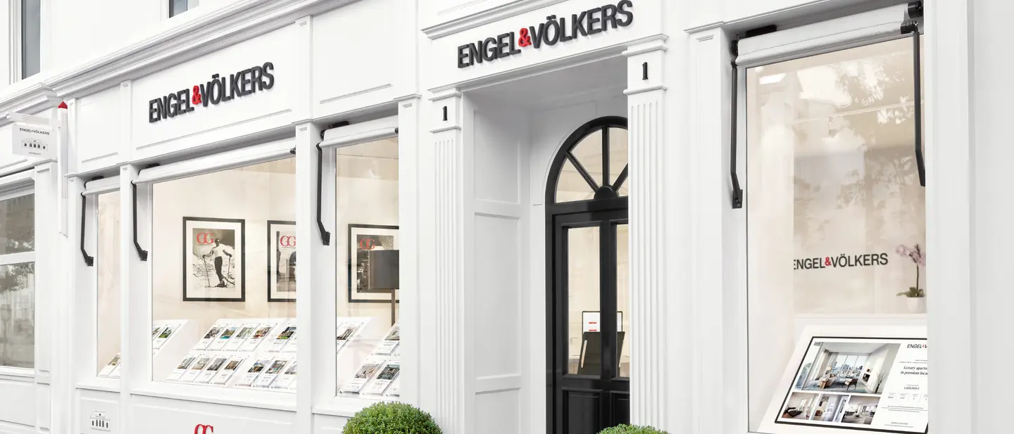 Engel & Völkers real estate office exterior. White building with black door and signage. Window displays feature property listings.