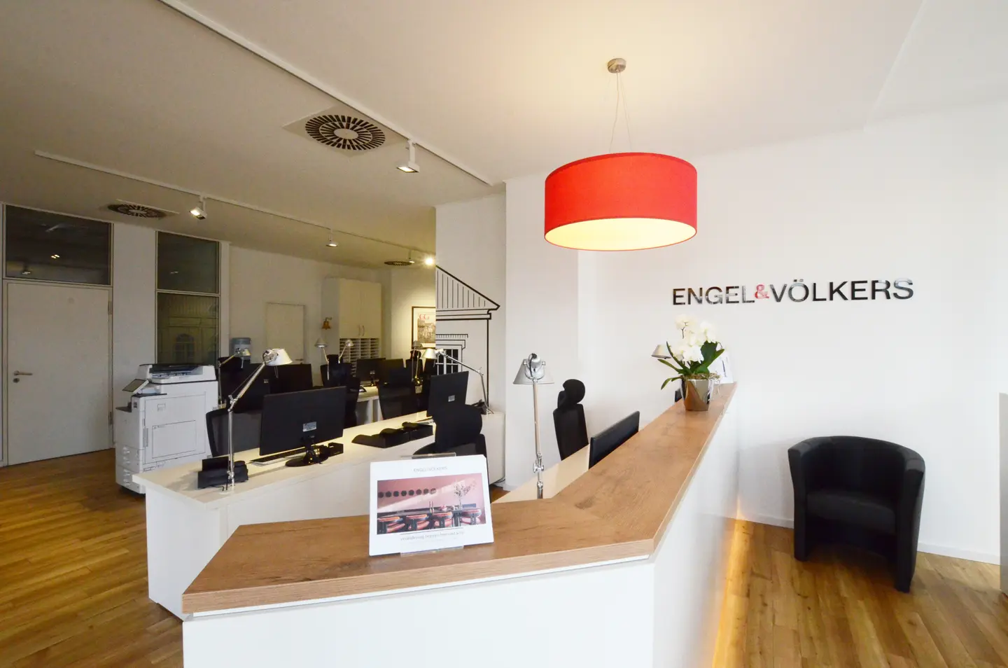 Engel & Völkers office reception area with a wood counter, white orchids, a red pendant light, and a black chair.