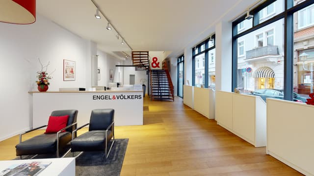 Engel & Völkers office interior with wood floors, white walls, black chairs, and a staircase with a red "&" symbol. Large windows show a street view.