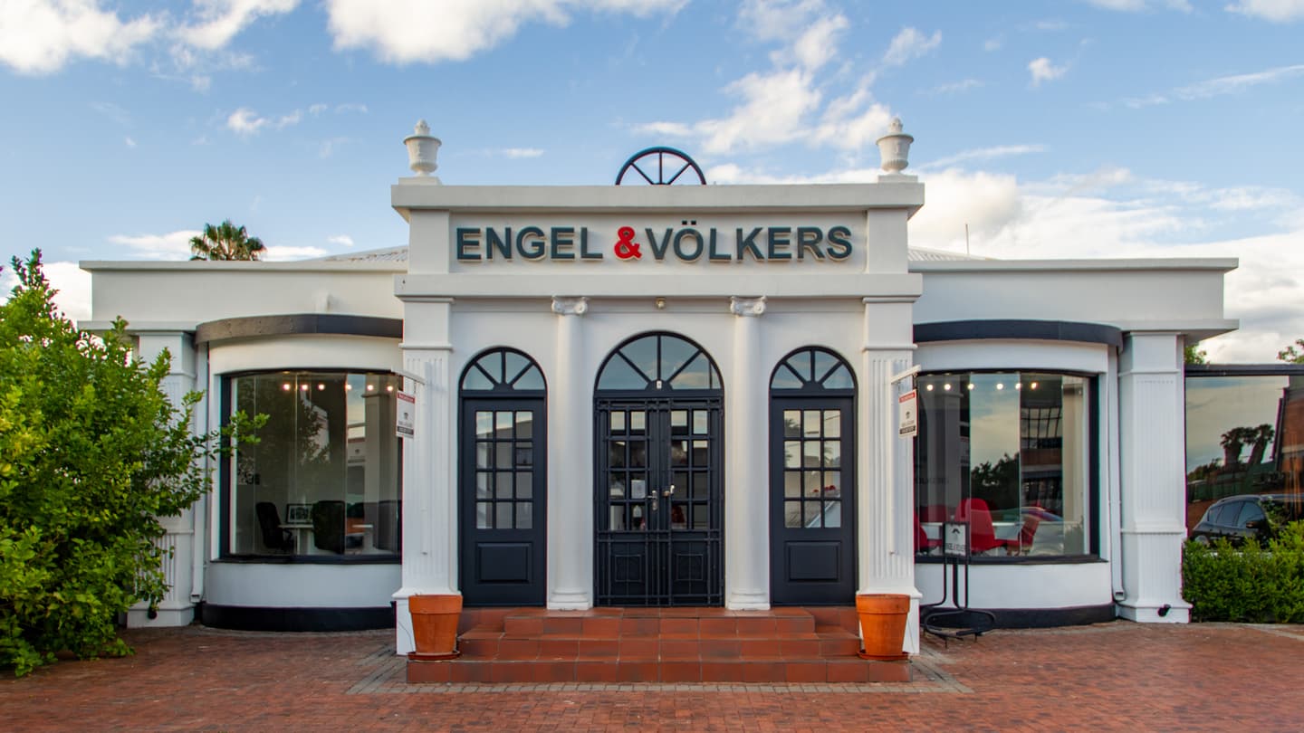 Engel & Völkers real estate office with white facade, black doors, and brick walkway under a cloudy sky.