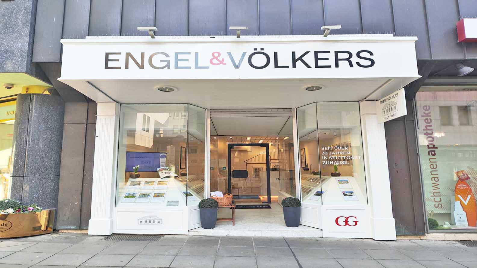 Engel & Völkers real estate storefront with glass windows displaying property listings. A basket sits on a small table near the entrance.
